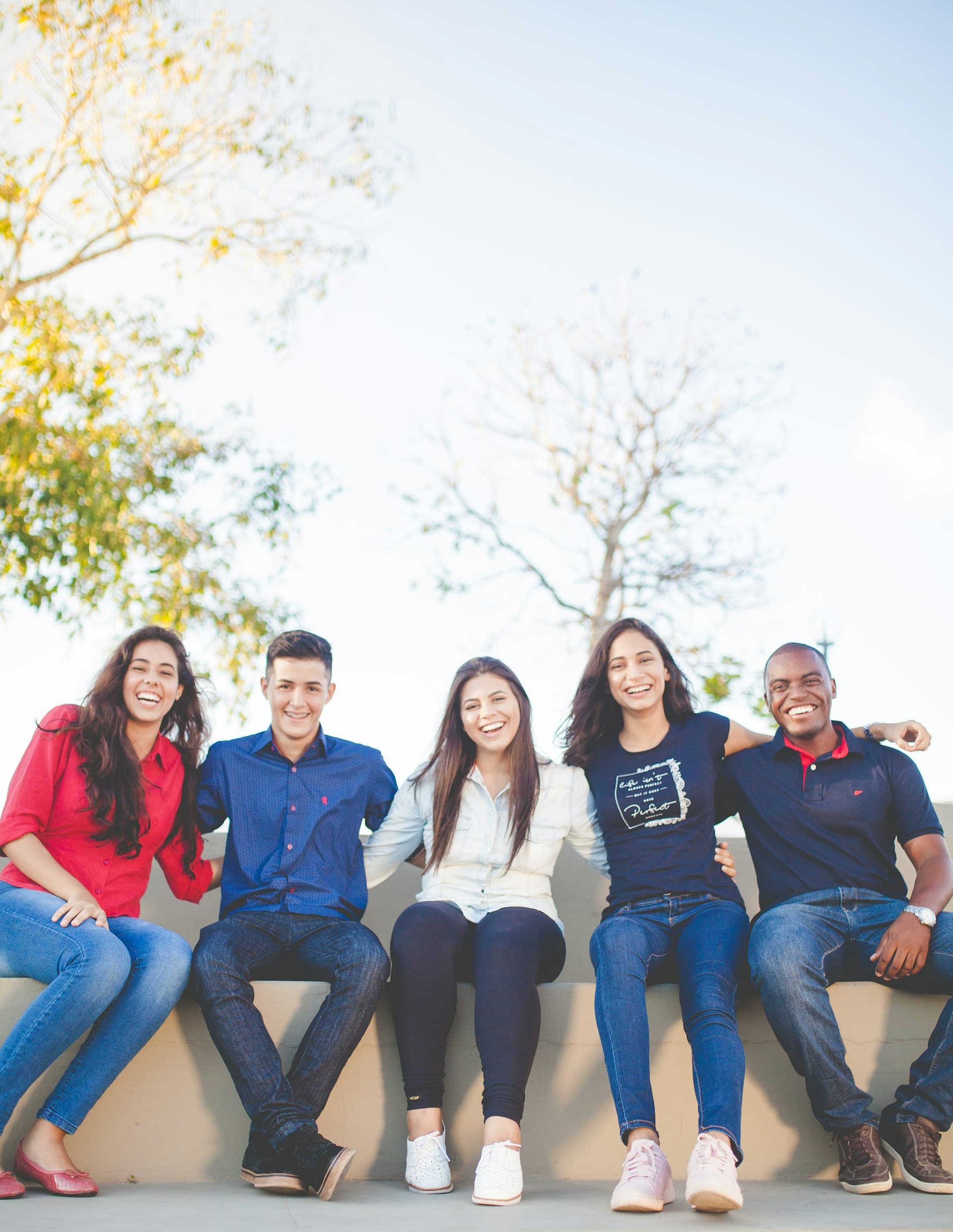 Five smiling teenagers sitting together outdoors with arms around each other